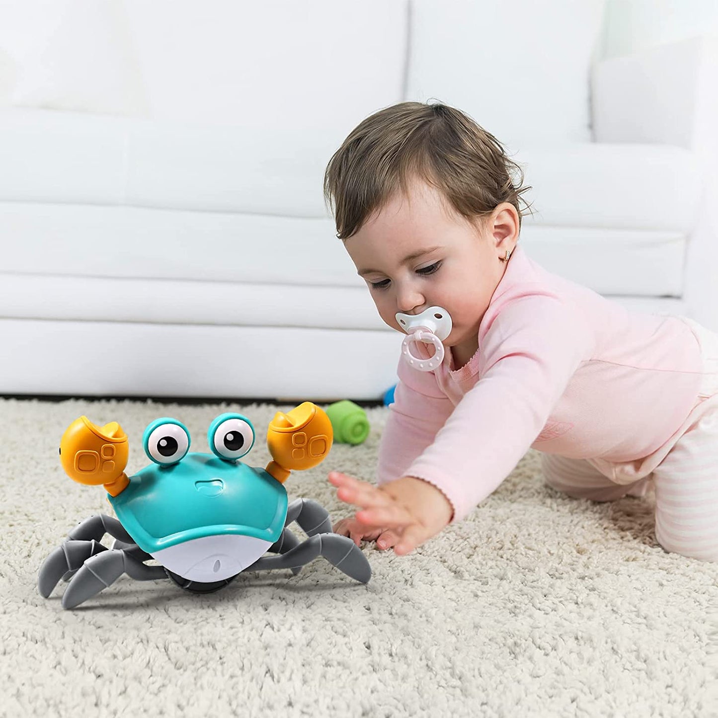 A baby in pink pajamas enjoys tummy time on a soft carpet, reaching for the Toyfetti HappyCrab – Crawling Companion with Lights & Music, a playful crab toy with big eyes and yellow claws. A pacifier is in the babys mouth beside a white sofa.
