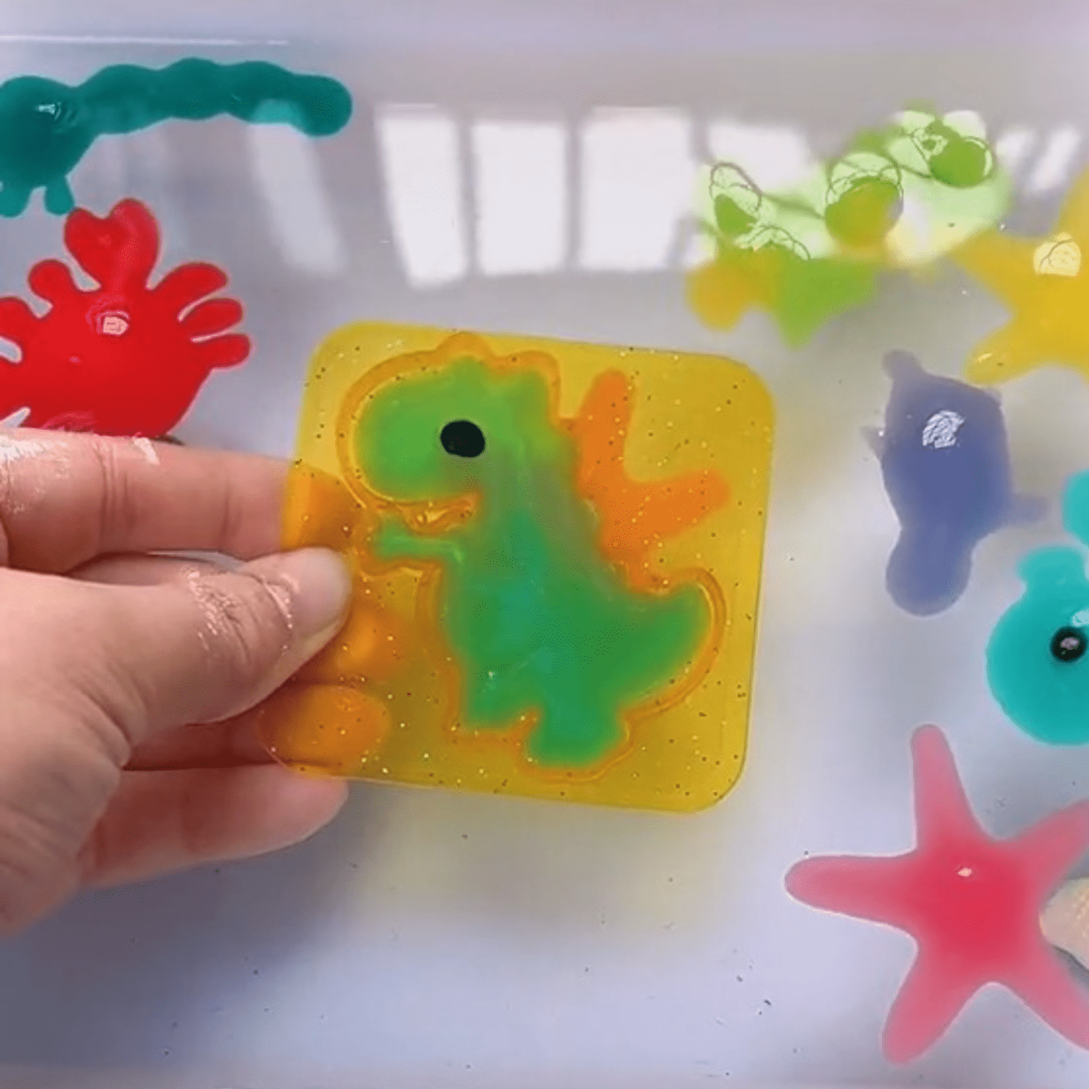 A hand displays a Toyfetti Magic Water Wonder Kit gel sticker featuring a green dinosaur, surrounded by vibrant 3D gel sea creatures and plants, perfect for sensory play and hands-on fun on a white surface.