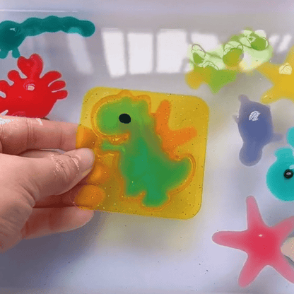 A hand displays a Toyfetti Magic Water Wonder Kit gel sticker featuring a green dinosaur, surrounded by vibrant 3D gel sea creatures and plants, perfect for sensory play and hands-on fun on a white surface.