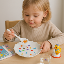 A young child sits at a table, using the Magic Water Pen – 12 Floating Drawing Markers above a white plate with colorful alphabet pasta. Beside them are markers, a yellow duck toy, and floating artworks, creating a joyful and focused scene.