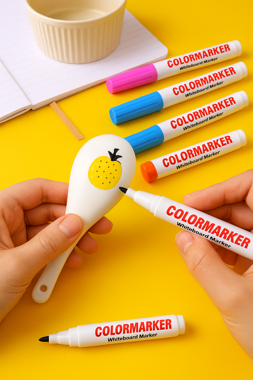 A person uses the Magic Water Pen – 12 Floating Drawing Markers to outline a white spoon with a yellow pinecone design. Six vibrant markers and a notebook lie on the yellow background, ready for more floating magic and doodles.