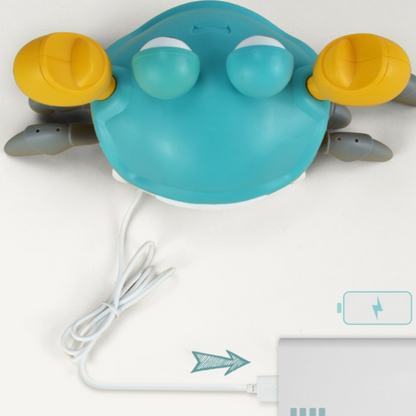 A blue and yellow Toyfetti HappyCrab – Crawling Companion with Lights & Music is connected to a white charging cable plugged into power, as an arrow and battery charging icon appear at the bottom right.