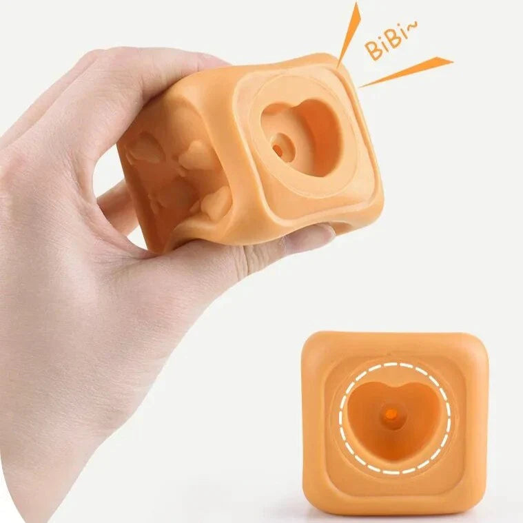 A hand squeezes a soft, orange CubbyKoala silicone teether & stacking toy by Toyfetti that makes a “BiBi~” sound, while another with a heart-shaped hole sits beneath. Both toys are cube-shaped and perfect for babies.