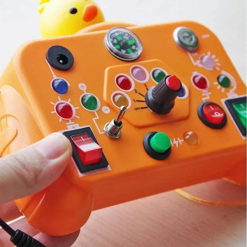 A hand presses a red switch on the Toyfetti BusyBoard Logic & Light Panel – Montessori Switch Board for Curious Kids, featuring an orange panel with colorful buttons, switches, a joystick, and a yellow duck; ideal for sensory play.