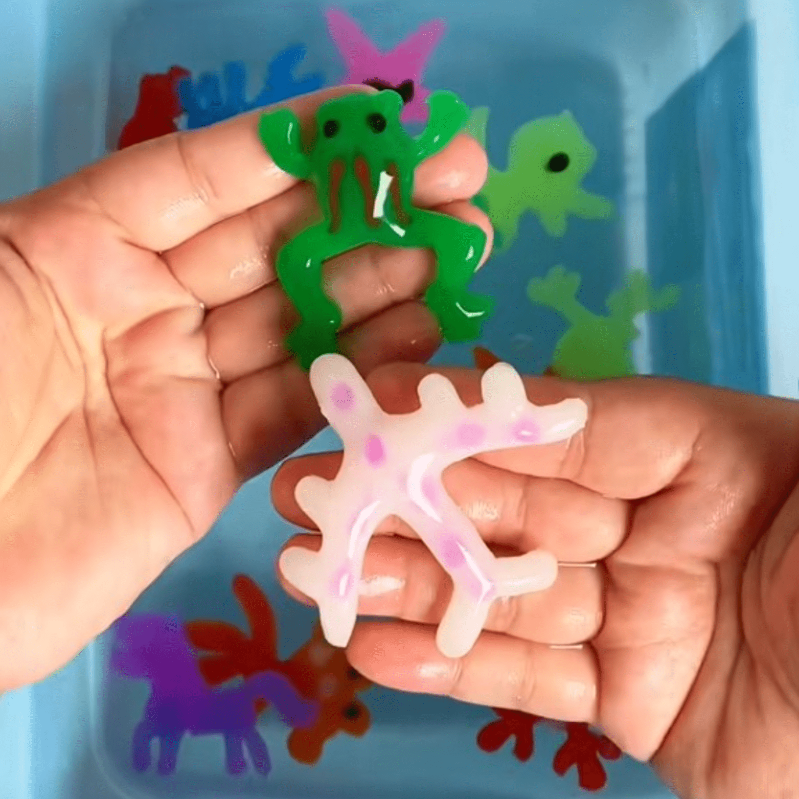 Two hands hold colorful, stretchy animal shapes—a green and a pink—over a container filled with more Toyfetti Magic Water Wonder Kit 3D animals. Their wet, translucent textures encourage sensory play and hands-on fun.