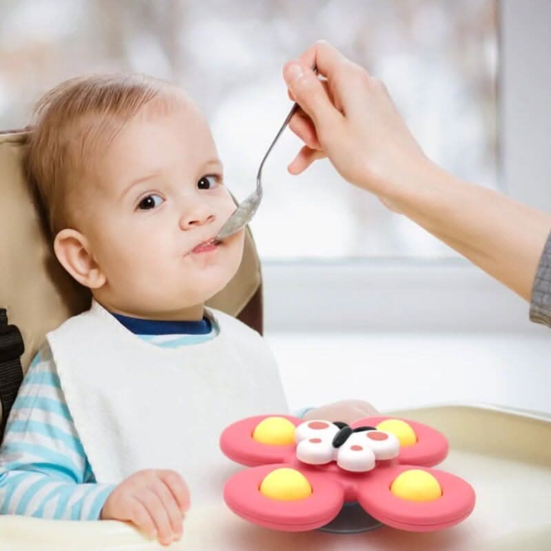 A baby in a high chair, wearing a white bib, is fed by an adult while the Toyfetti Whirlie Baby Spinner – Sensory Spin Toy for Fine Motor Fun sits on the tray in front of them.