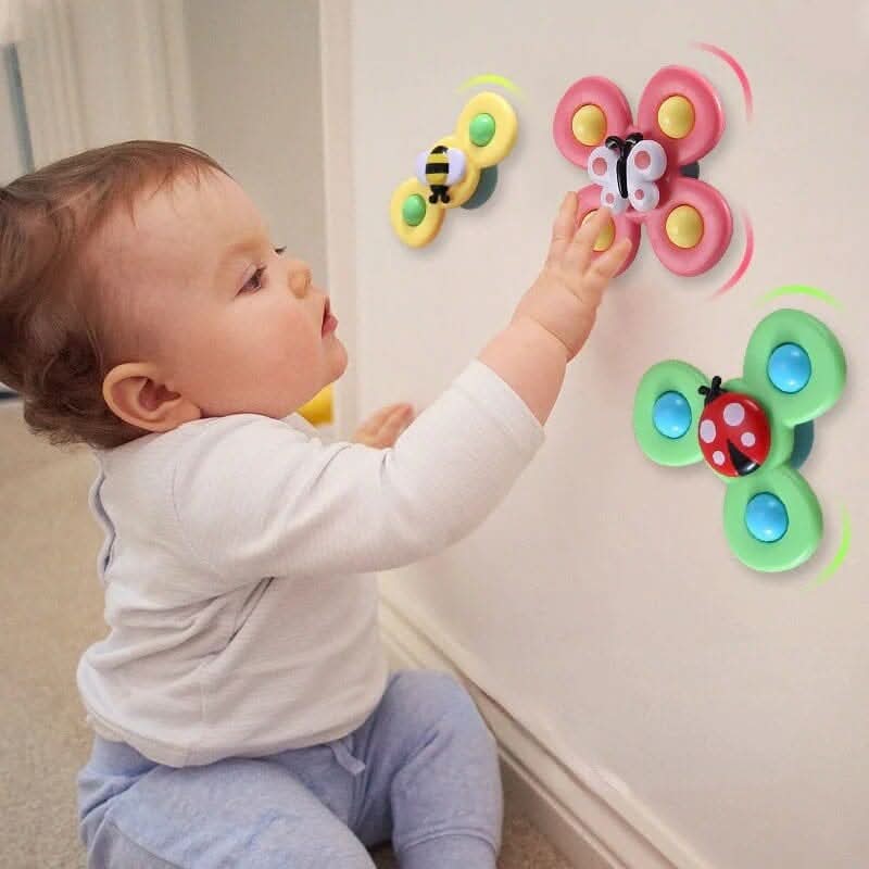 A baby in a white shirt and blue pants sits on the floor, reaching for Toyfettis Whirlie Baby Spinner – Sensory Spin Toy for Fine Motor Fun, colorful insect-themed spinners that promote sensory development as they spin on the wall.