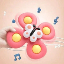 A hand holds the Toyfetti Whirlie Baby Spinner, a pink butterfly-shaped sensory spin toy with yellow wing circles, against a peach background with music notes and stars, suggesting it plays sounds to support fine motor skill development.