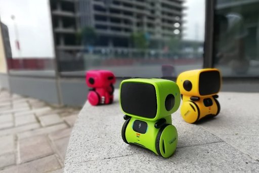 Three Toyfetti Robomate | Jouw Muzikale Vriend - Slimme Robot toys in green, pink, and yellow are outdoors on a stone surface, with a blurred cityscape and buildings visible in the background.
