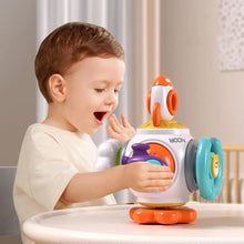 A young child with short brown hair smiles while playing at a table with the Toyfetti Montessori Sensory Activity Cube – Press, Turn & Explore Learning Toy for Toddlers.