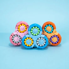 Five BrainBounce Rotating Logic Spinner toys by Toyfetti, each in pink, blue, orange, and purple with circular designs and beads, are arranged on a blue surface against a light blue background.