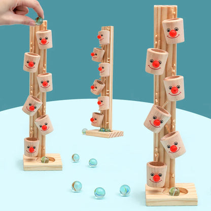 Three Toyfetti Clown Track Wooden Play Towers with smiling faces and red noses are shown. A hand drops a marble in, promoting fine motor skills and imaginative play. Blue-green marbles are scattered on a light blue surface.