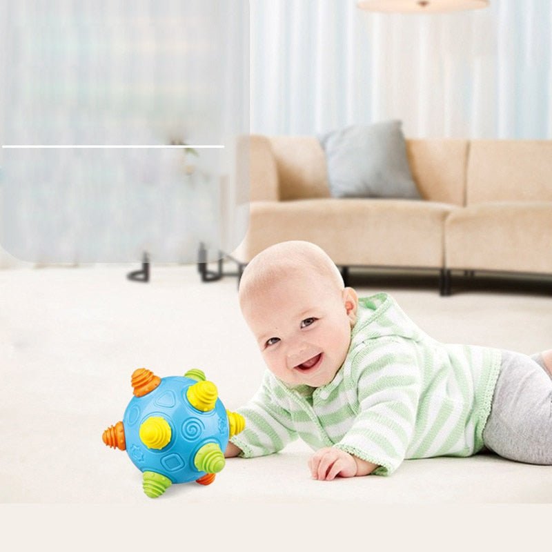 A smiling baby in a light green striped hoodie lies on a white carpet, reaching for the Toyfetti Sensory Music Ball – Interactive Light & Sound Toy for Babies and Toddlers. A beige sofa with a cushion is visible in the background.