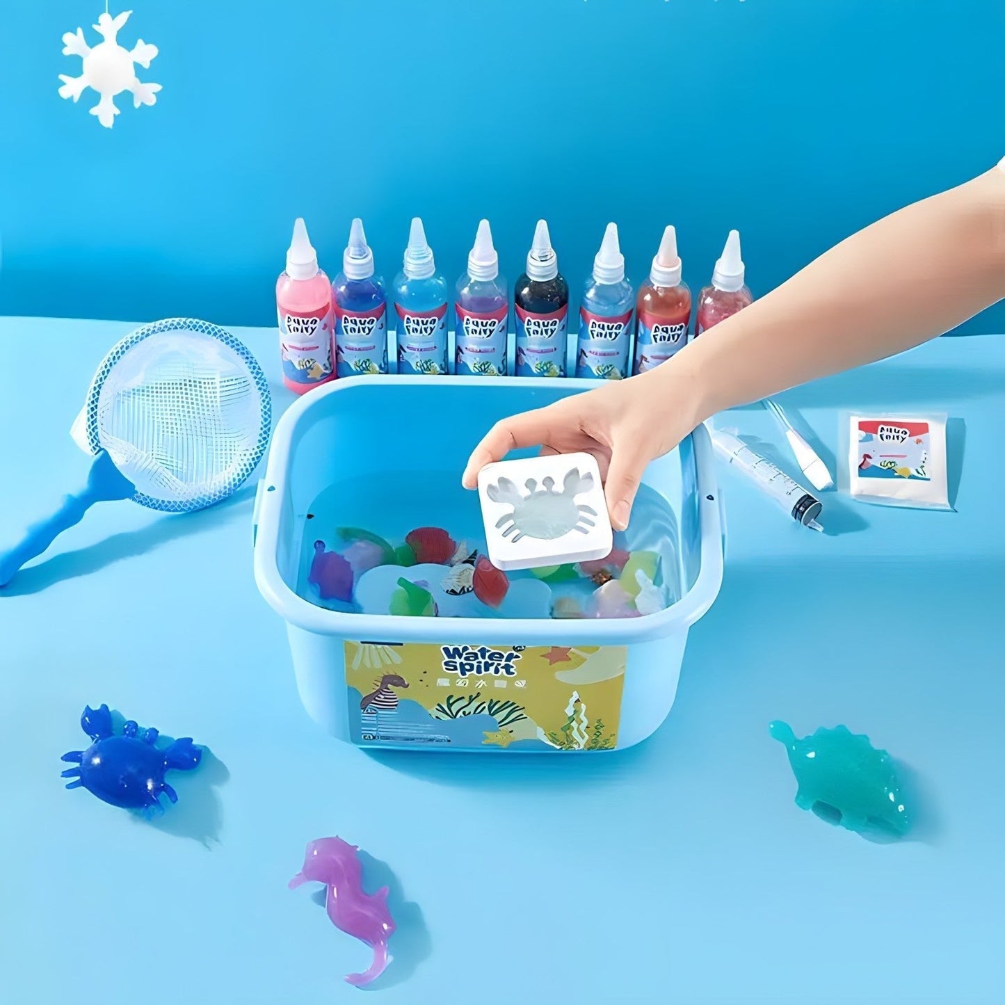 A hand dips a white crab mold from the Toyfetti Magic Water Wonder Kit – Create Colorful 3D Animals with Just Water into a blue tub, surrounded by bottles, nets, and crafting supplies on a table with colorful sea creatures and a blue backdrop.